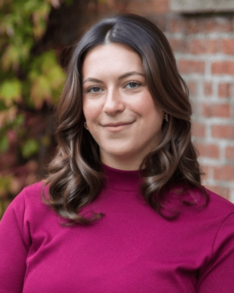 Portrait of a woman with shoulder-length brown hair wearing a maroon sweater, standing outdoors in front of a brick wall and fall foliage.